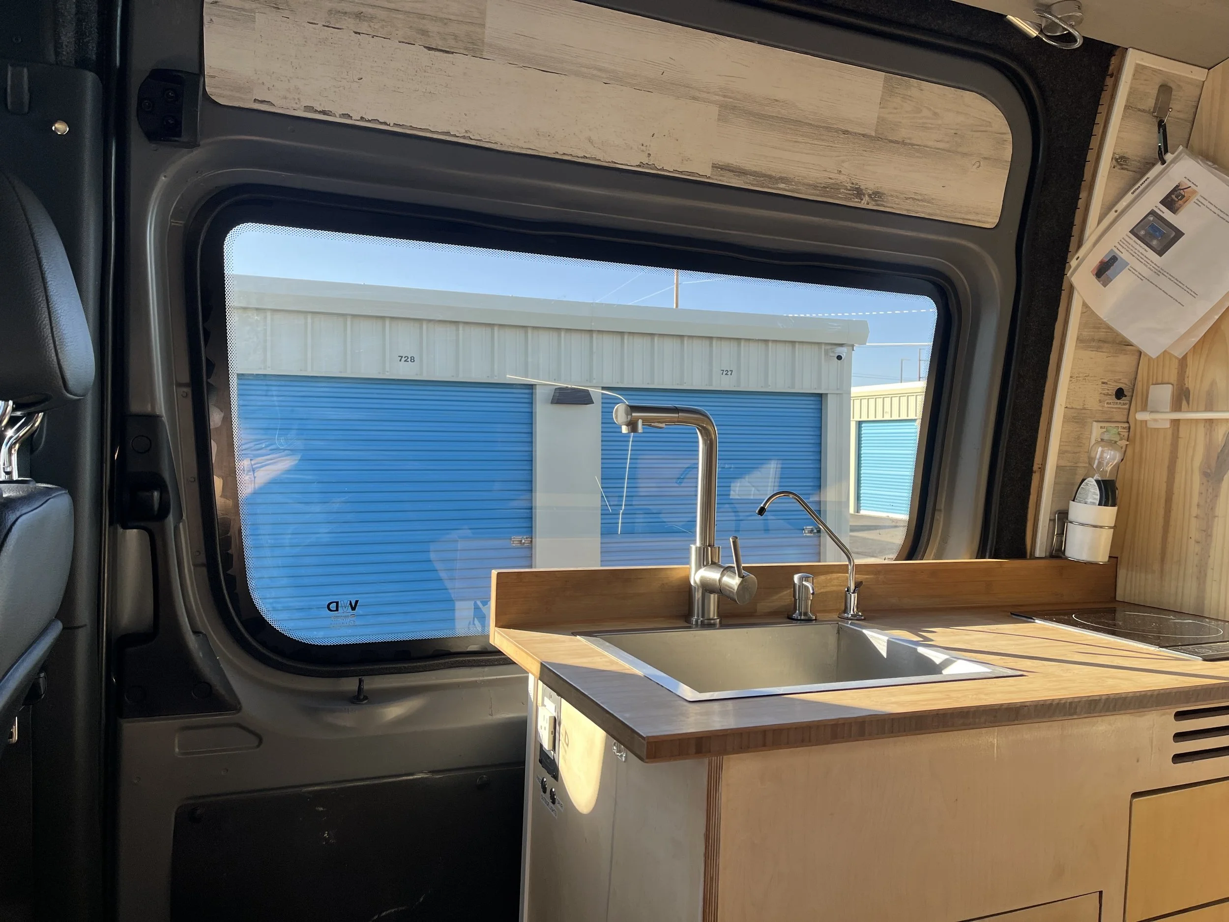 Interior of a tiny home or camper with a kitchen area, featuring a stainless steel sink, faucet, and a countertop with a stove, situated near a large window with a view of storage units.