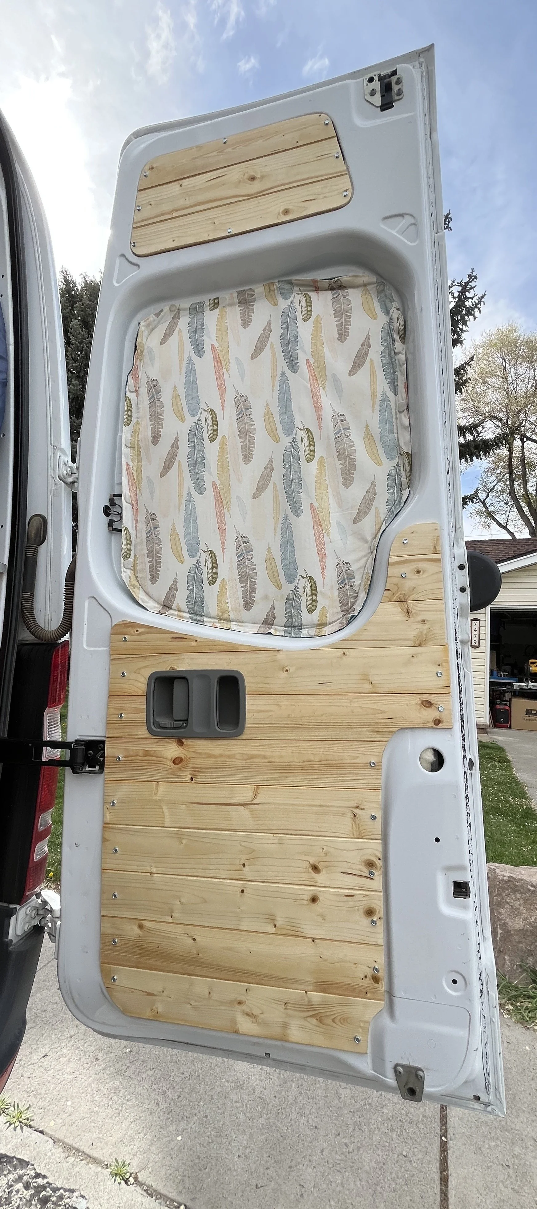 The inside of a van showing a wood-paneled wall, a window with a fabric curtain with feathers, and a small storage compartment.