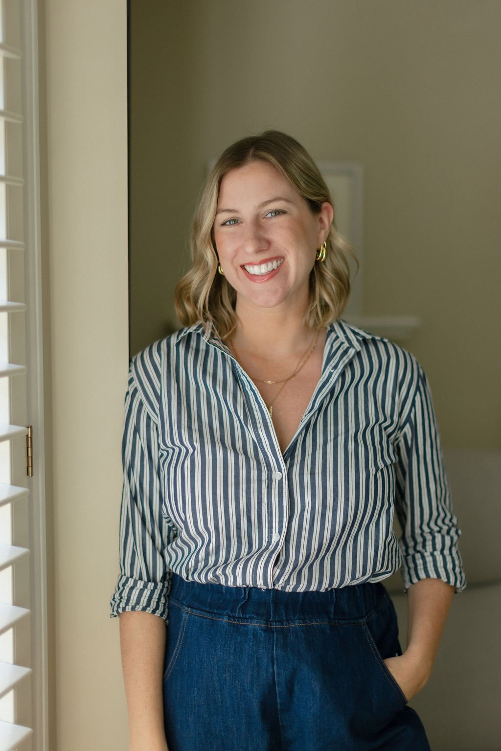 Smiling woman with shoulder-length blond hair, wearing a striped button-up shirt and blue jeans, standing indoors near a window with white shutters.