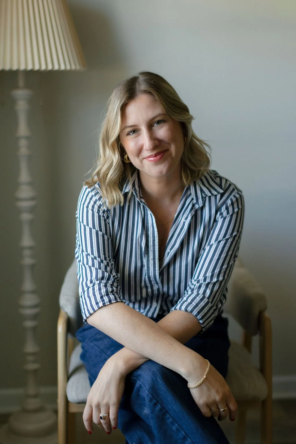 A woman with shoulder-length blonde hair, wearing a blue and white striped shirt and blue jeans, sitting on a small wooden chair with her arms crossed and smiling at the camera. There is a beige lamp in the background on the left side of the image.