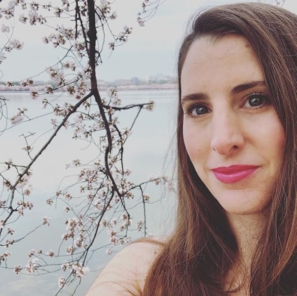 A woman with long brown hair and pink lipstick takes a selfie outdoors near a body of water with cherry blossom branches in the background.