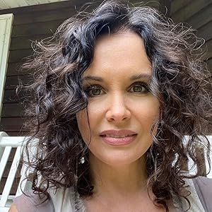 Close-up of a woman with curly gray hair and light makeup, outdoors near a wooden structure.