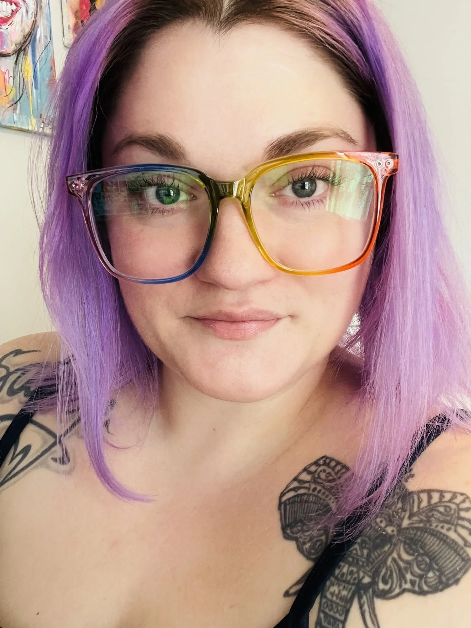 Close-up of a woman with purple hair wearing multi-colored glasses, showing her face, tattoos on her shoulder and chest, and part of her shirt.