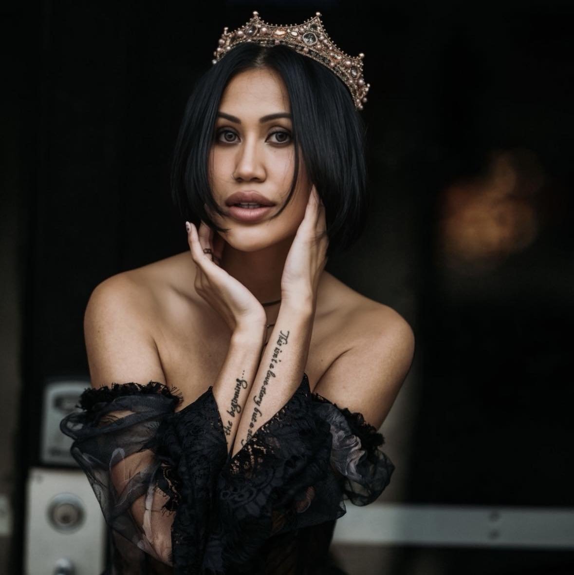 A woman with short black hair wearing a pearl and gemstone crown, dressed in black lace off-the-shoulder clothing, holding her face with both hands, against a dark background.