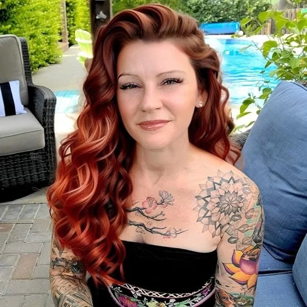 A woman with long, wavy red hair and tattoos on her shoulders and arms is sitting outdoors near a swimming pool, smiling at the camera.