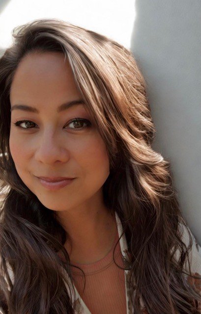 Close-up of a woman with long, wavy brown hair and a slight smile, posing next to a light gray wall.
