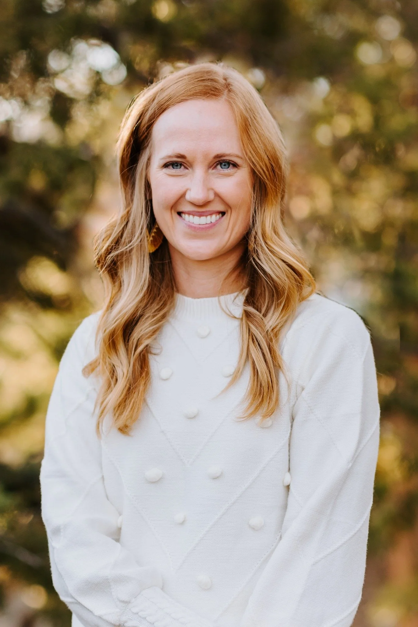 A woman with long, wavy red hair and blue eyes smiling outdoors, wearing a white sweater with decorative buttons, with a blurred natural background of trees and sunlight.