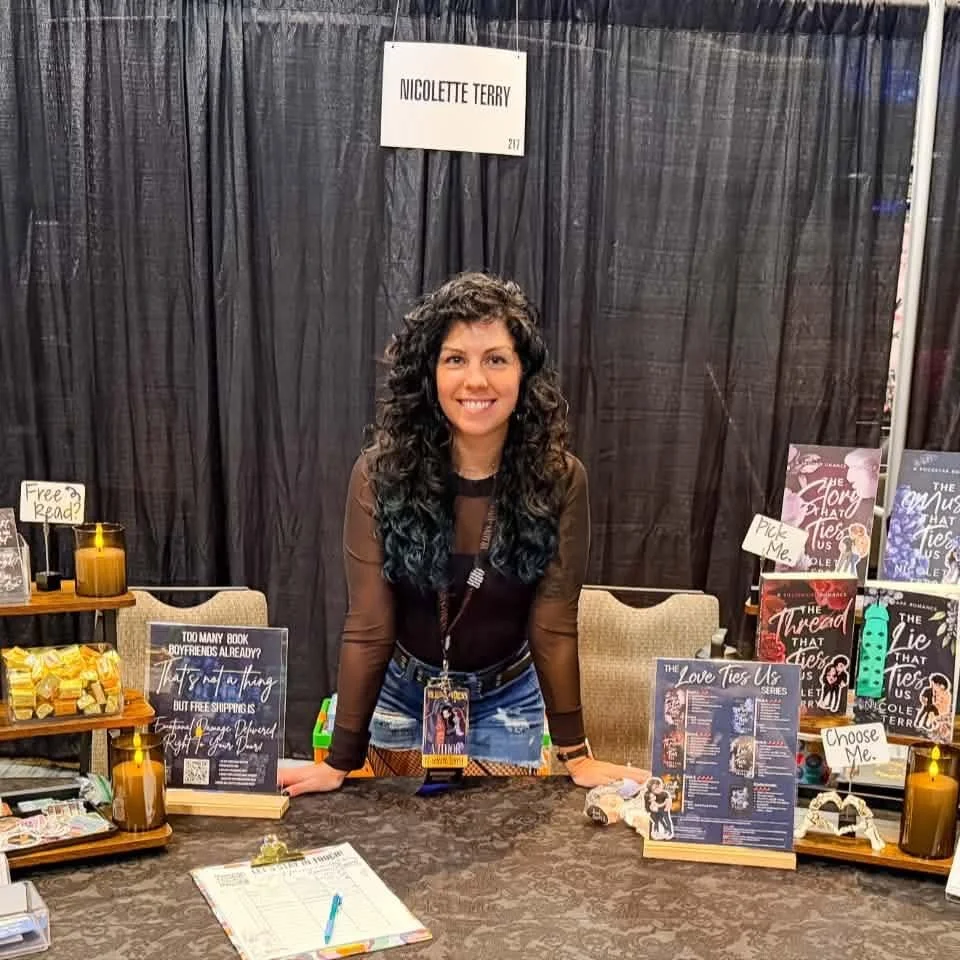 A woman with curly dark hair, wearing a brown sheer long-sleeve top and denim shorts, is standing behind a table at a book signing event. The table displays books, signs, and decorative items. A sign above her reads 'Nicolette Terry'.