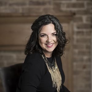 A woman with dark curly hair smiling, wearing a black top and a gold necklace, sitting against a brick wall background.