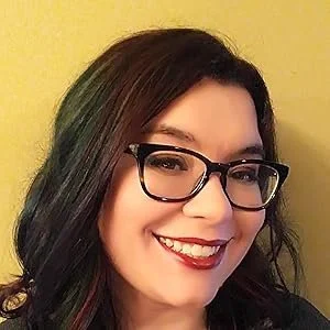 A woman with black hair and glasses smiling in front of a yellow wall.
