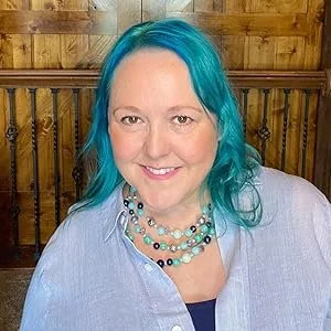 A woman with bright blue hair smiling, wearing a light-colored shirt and a layered beaded necklace, in front of a wooden background.