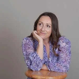 A woman with long brown hair resting her chin on her hand, wearing a purple floral blouse, sitting on a wooden chair against a plain gray background.