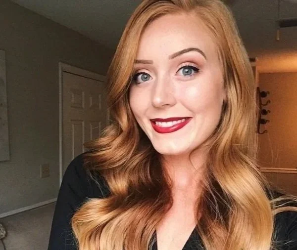 A young woman with wavy red hair and bright makeup smiling in a home setting.