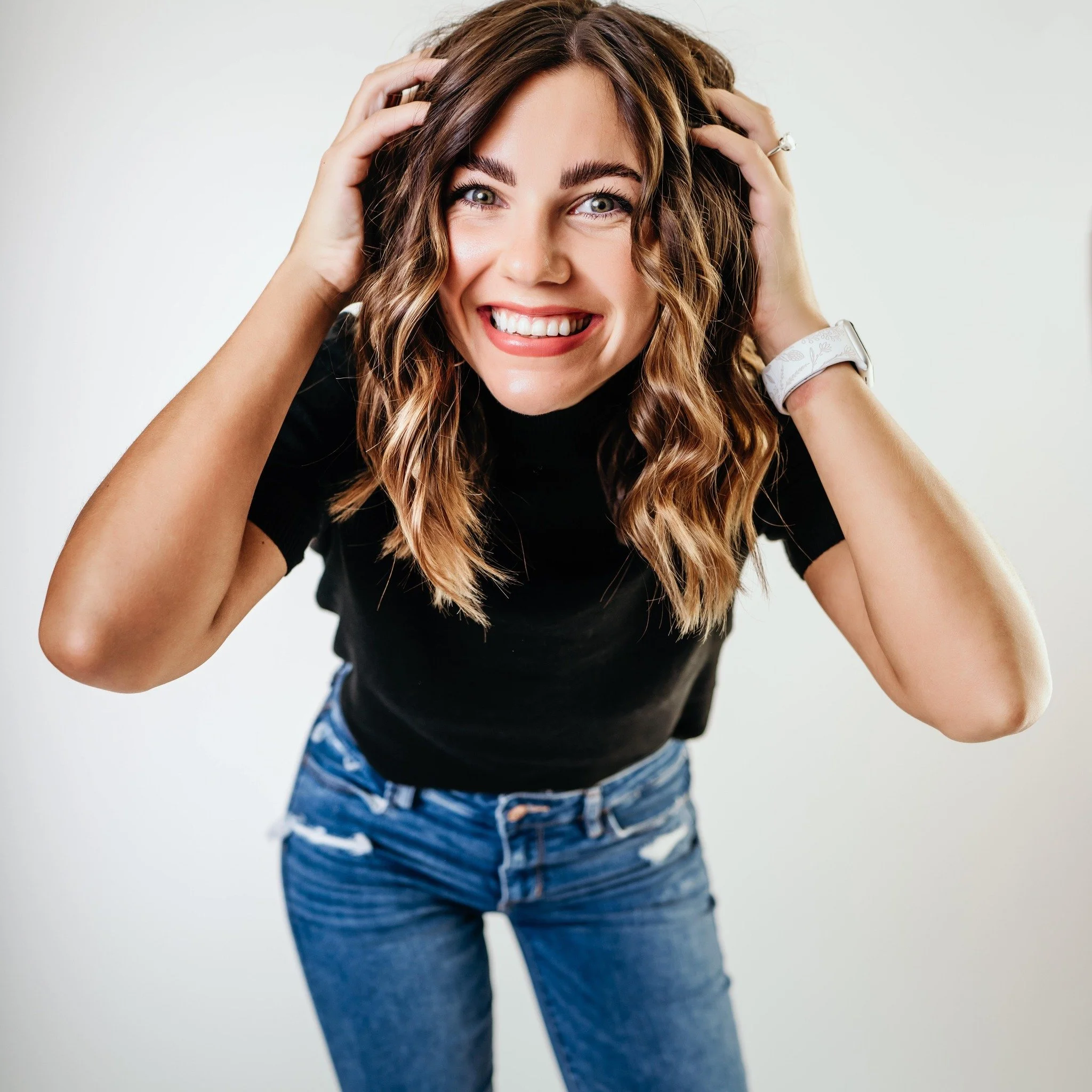 Close-up of a smiling woman with wavy light brown hair, wearing a black top and blue jeans, holding her hair with both hands against a plain white background.