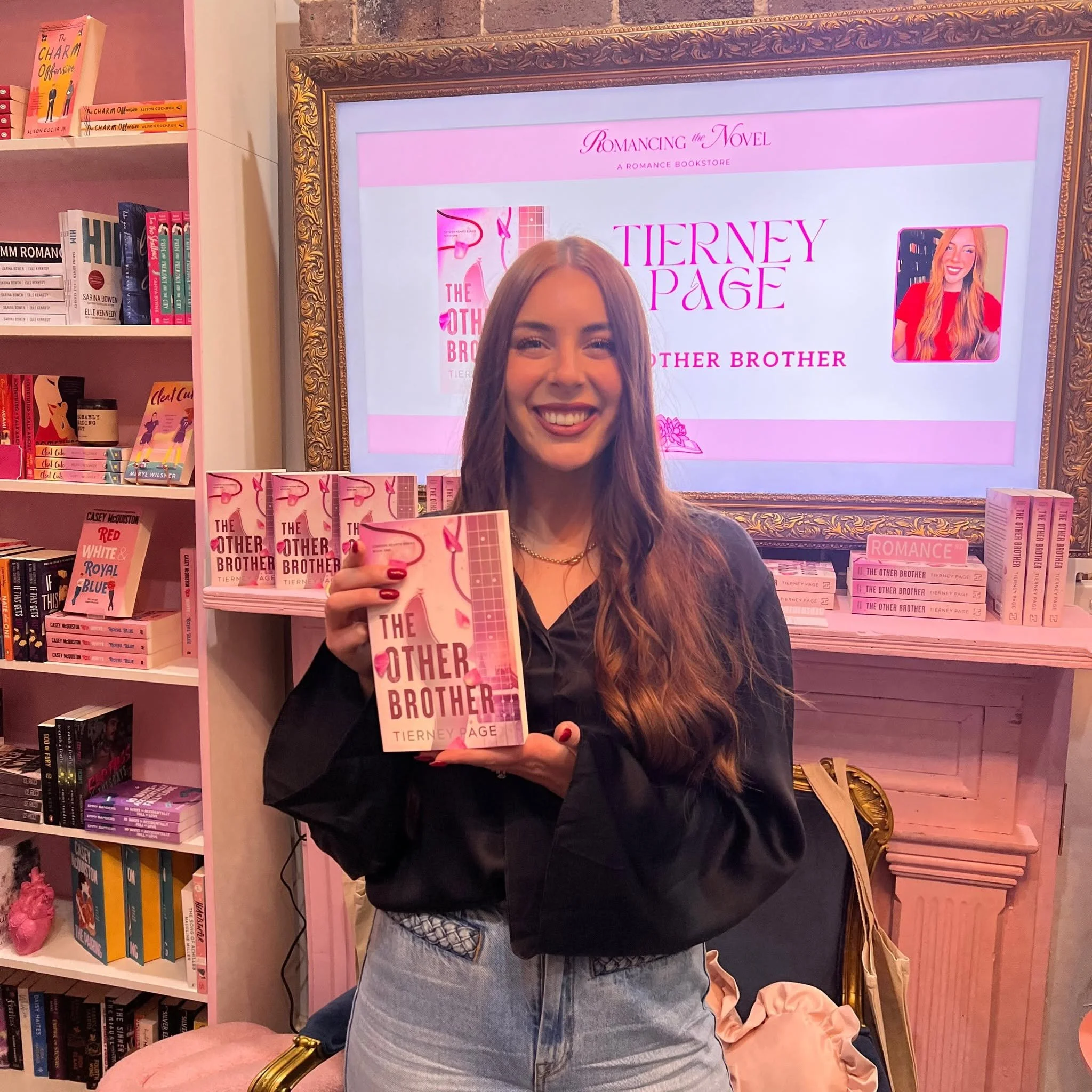 A young woman with long, wavy brown hair smiling while holding a pink copy of the book "The Other Brother" by Tierney Page at a bookstore. Behind her is a bookshelf filled with books, and a digital display advertising the same book on the wall.