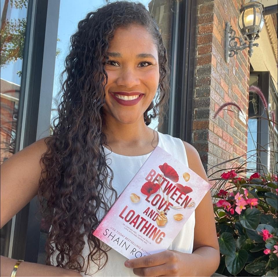 Woman with long curly hair holding a book titled 'Between Love and Loathing' outside near a brick wall and pink flowers.