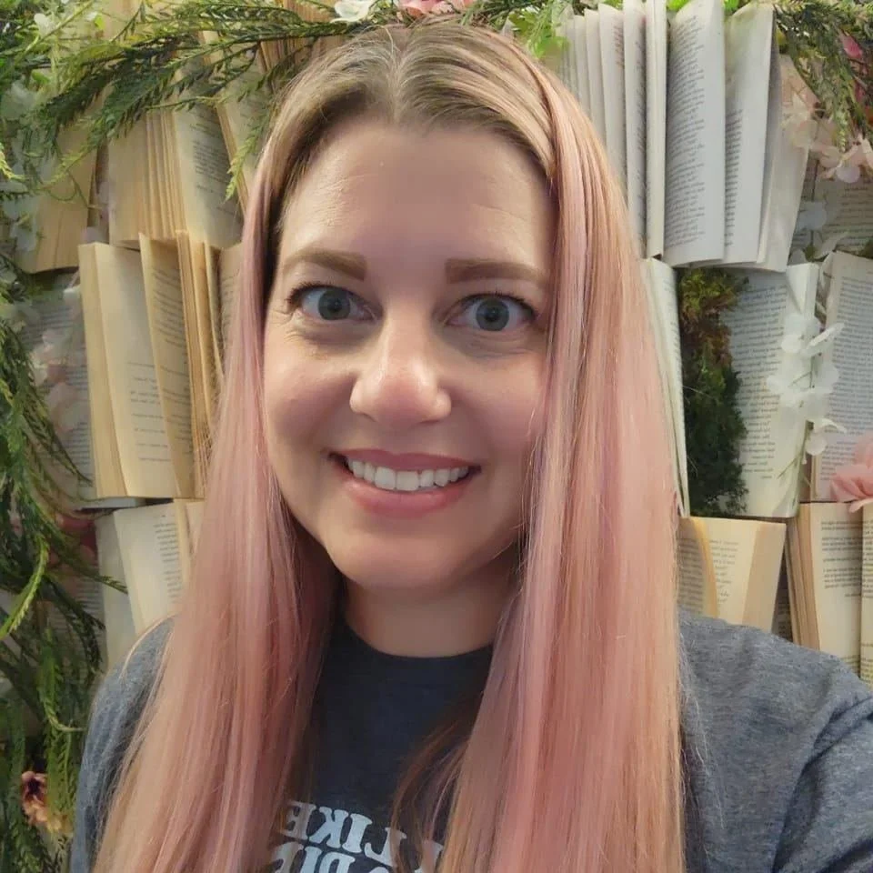 A woman with long pink hair and blue eyes smiling, standing in front of a decorative background made of open books and greenery.
