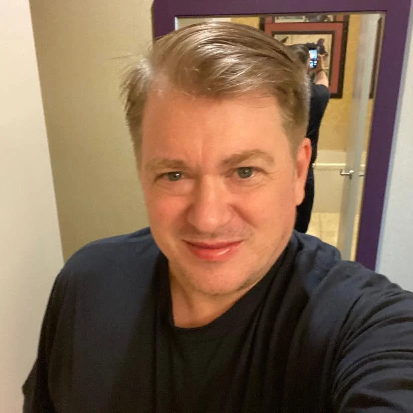 A man taking a selfie in front of a mirror in a room with beige and purple walls, with framed pictures on the wall behind him.