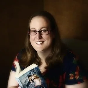 A woman with long hair and glasses holding a book, smiling in a dimly lit room.
