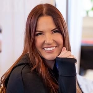 A woman with red hair smiling and resting her chin on her hand in an indoor setting.