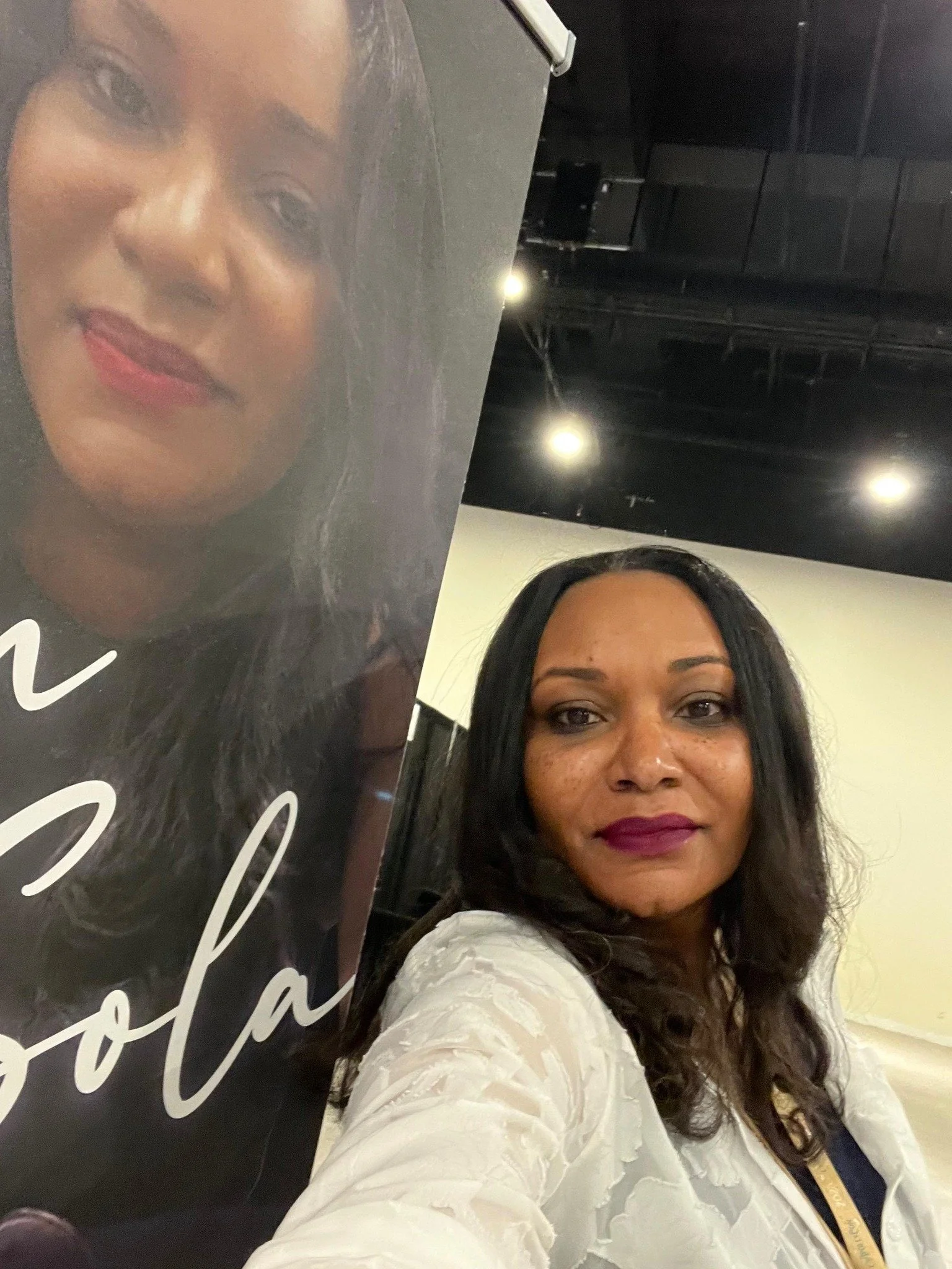 A woman with dark hair and brown skin taking a selfie next to a poster of another woman with similar features. The woman is wearing a white textured top and has a purple lipstick.