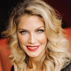 Close-up of a woman with long, wavy blonde hair, blue eyes, and red lipstick, smiling at the camera.