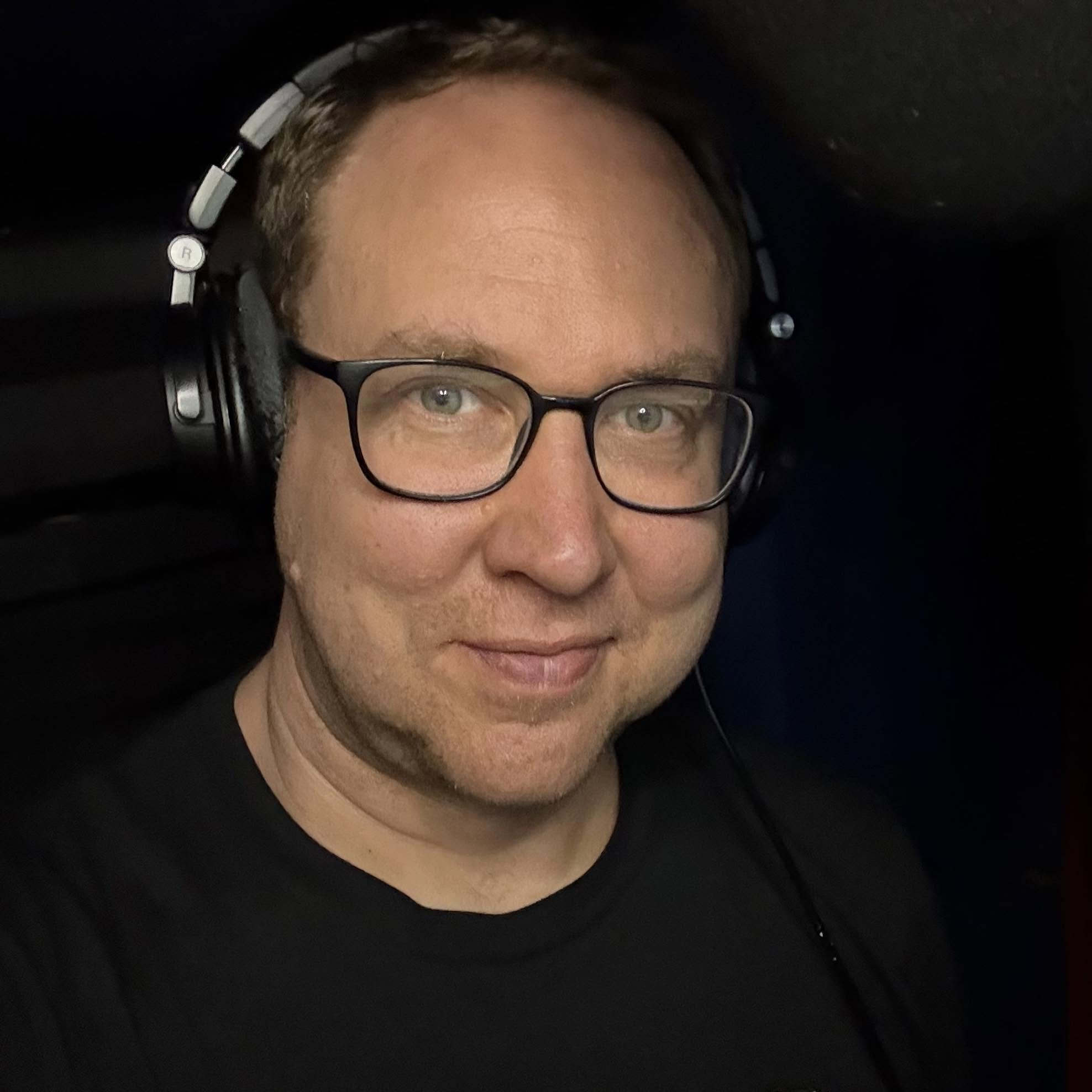 A close-up photo of a man with glasses and short hair, wearing headphones, partially visible with a dark background.