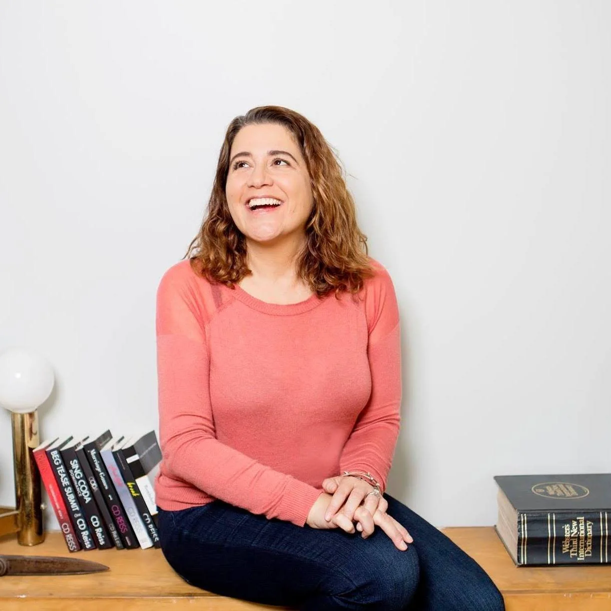 A woman with curly brown hair wearing a coral long sleeve shirt and dark jeans sitting on a wooden table, smiling and looking up. There are books and a white lamp next to her against a white wall.