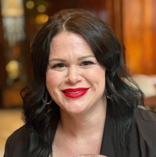 A woman with dark wavy hair and red lipstick, smiling in a warmly lit indoor setting.