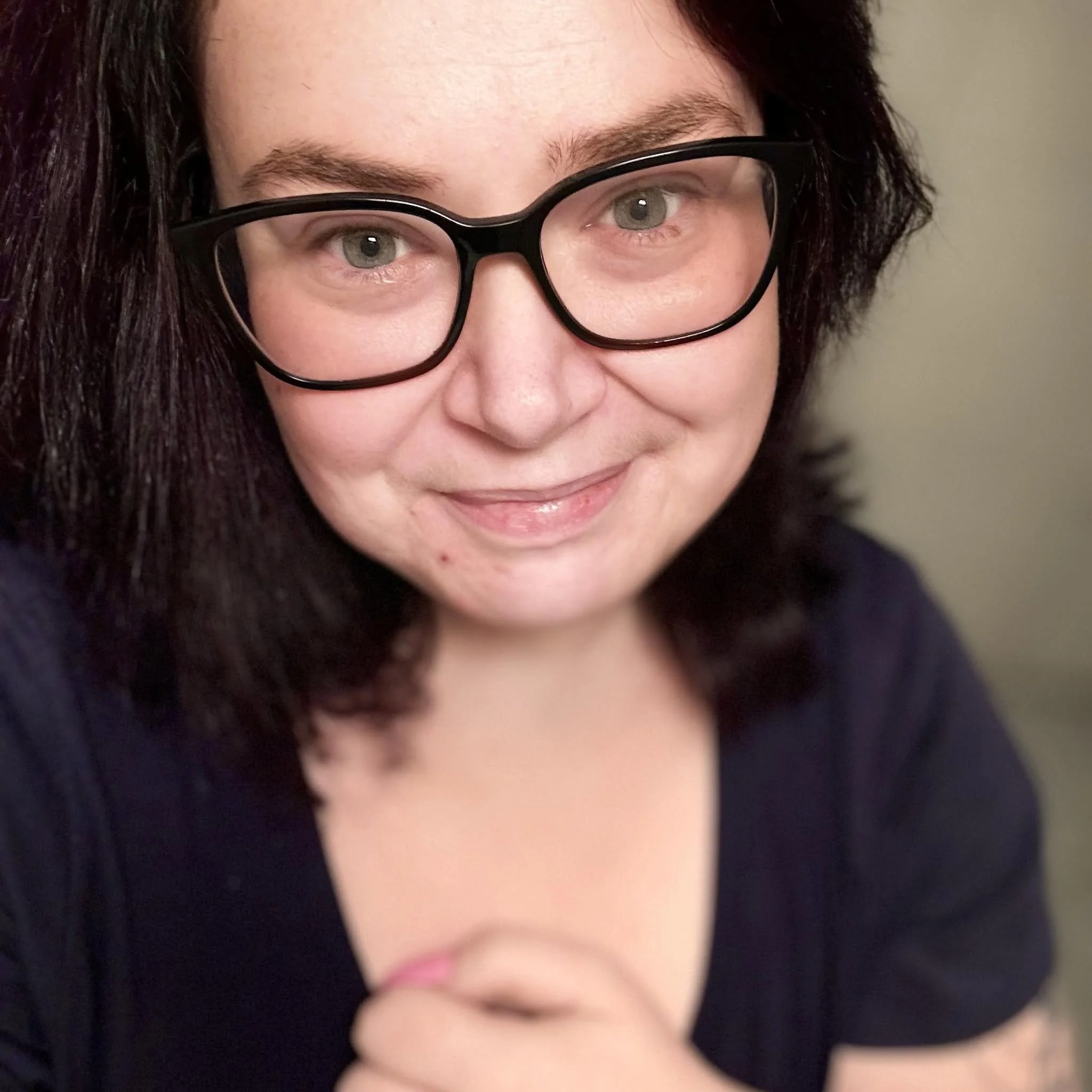 Close-up of a woman with light skin, glasses, and dark hair, smiling with her lips slightly parted.