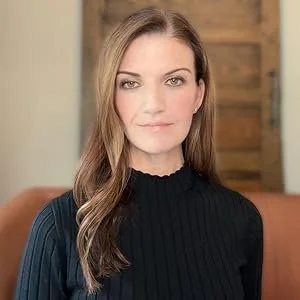A woman with long brown hair and fair skin, wearing a black ribbed turtleneck, posing in front of a wooden background.