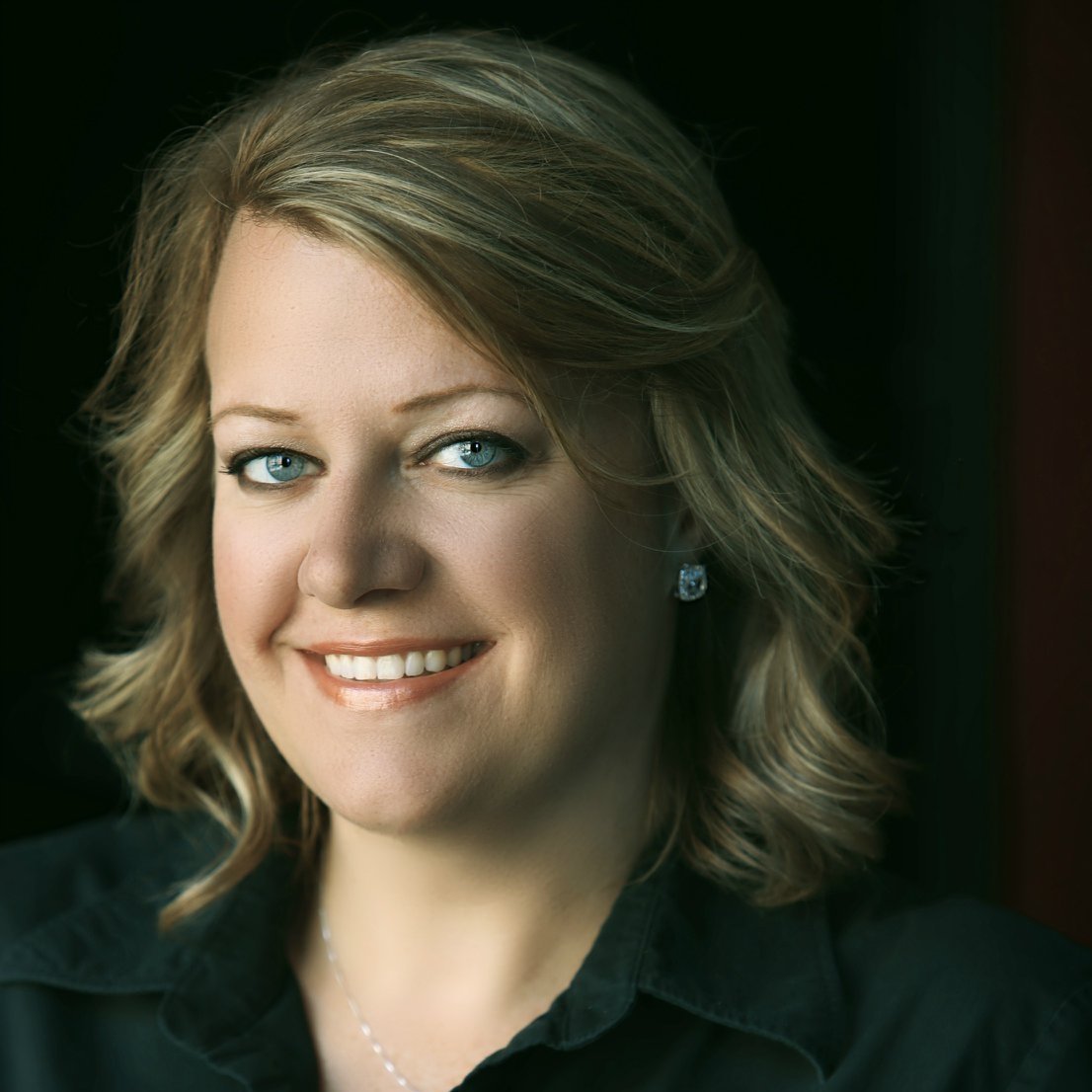 A portrait of a woman with blonde hair, blue eyes, smiling, wearing earrings and a dark top, against a dark background.