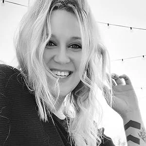 A young woman with blonde hair smiling at the camera, wearing a dark sweater, with string lights in the background.