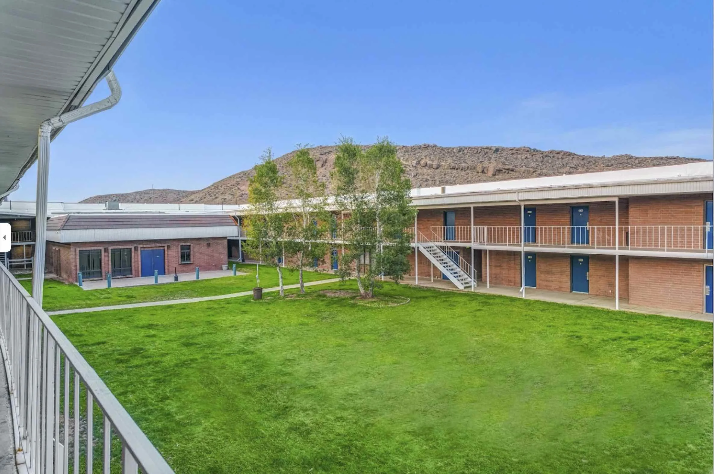 View from a balcony showing a courtyard with green grass, three trees, and a two-story building with red brick walls and blue doors. Hill in the background under a clear blue sky.