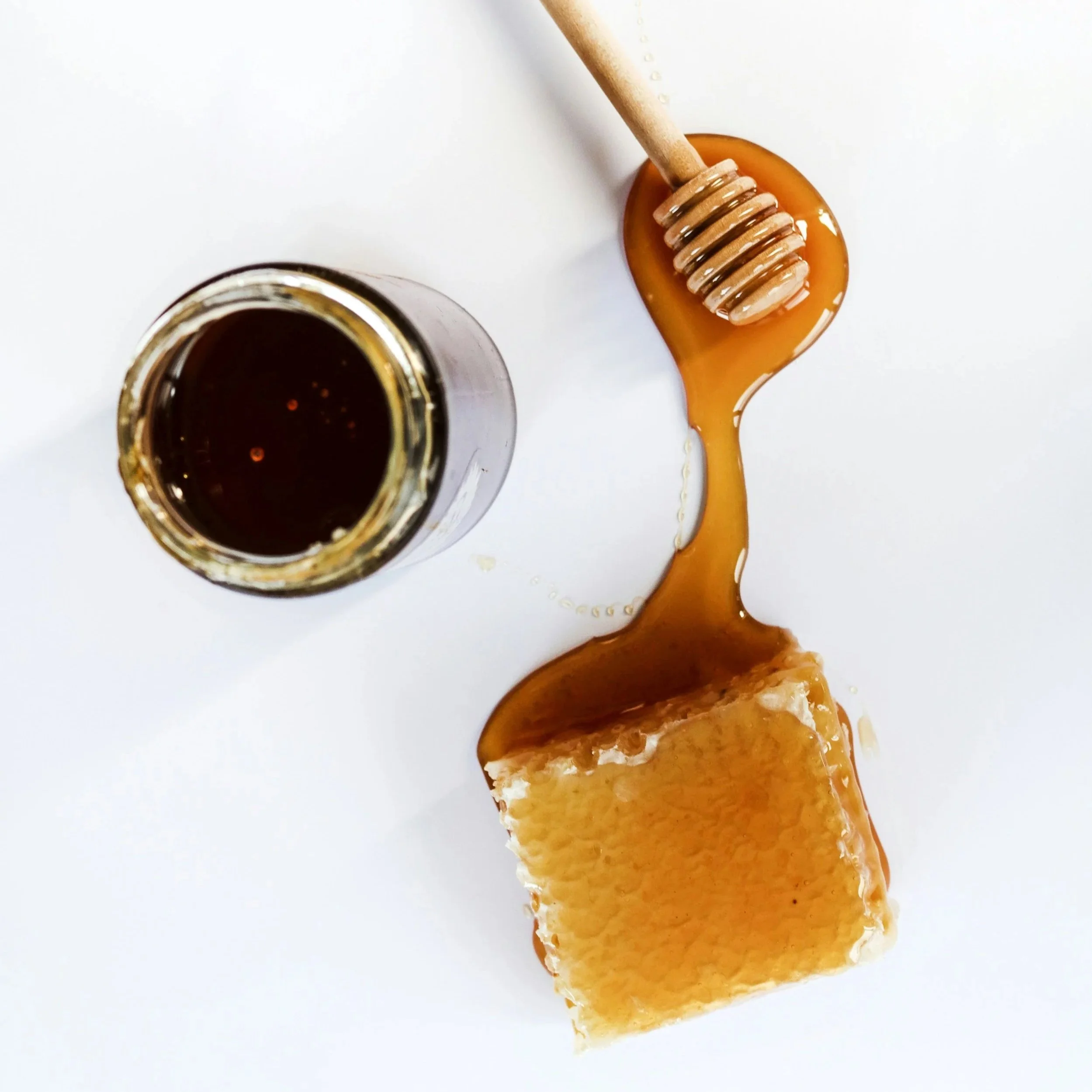 Wooden honey dipper drizzling honey onto a honeycomb beside an open jar of honey.