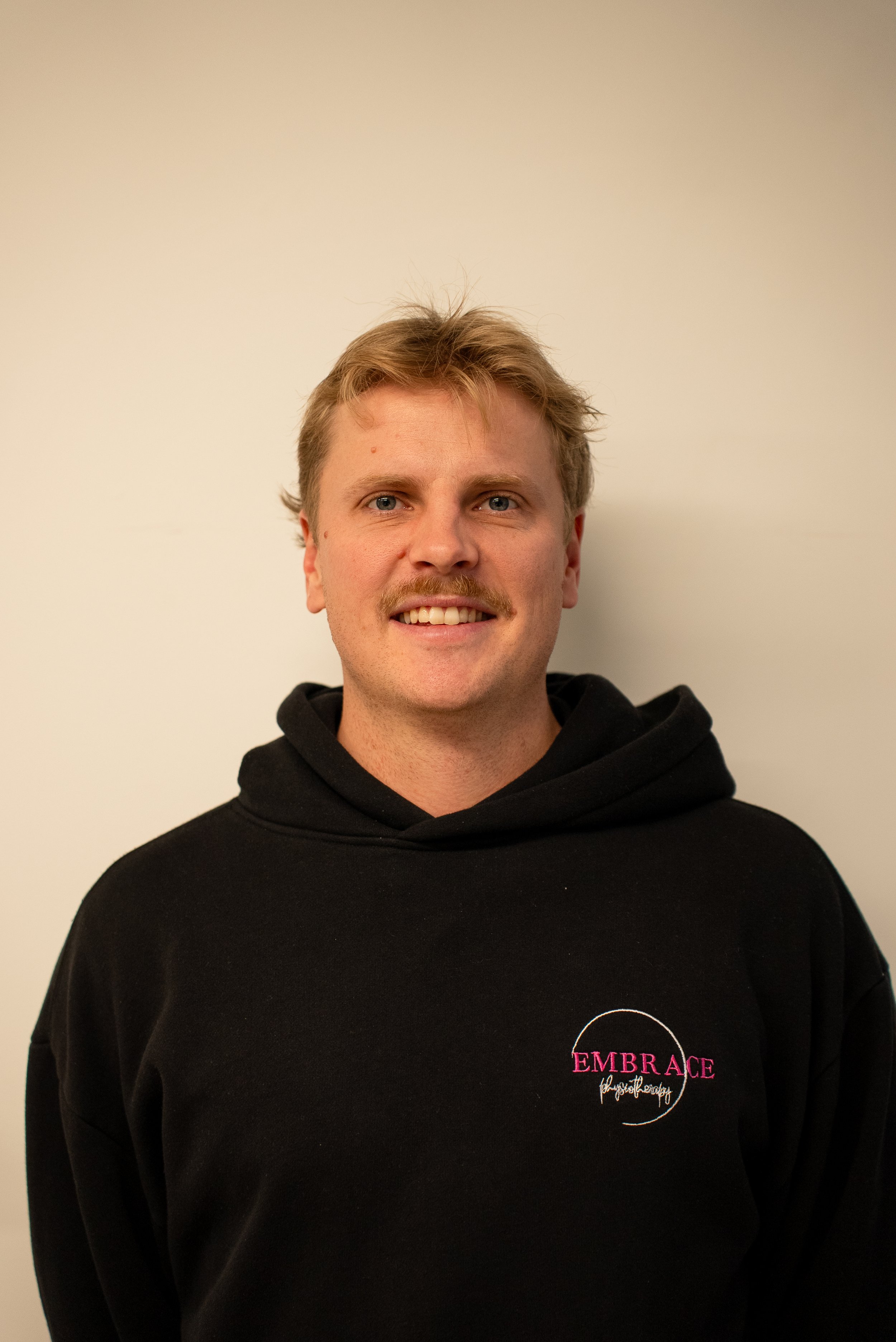 Young man with blonde hair and a mustache smiling, wearing a black hoodie with Embrace Physiotherapy. Physio Burnside 