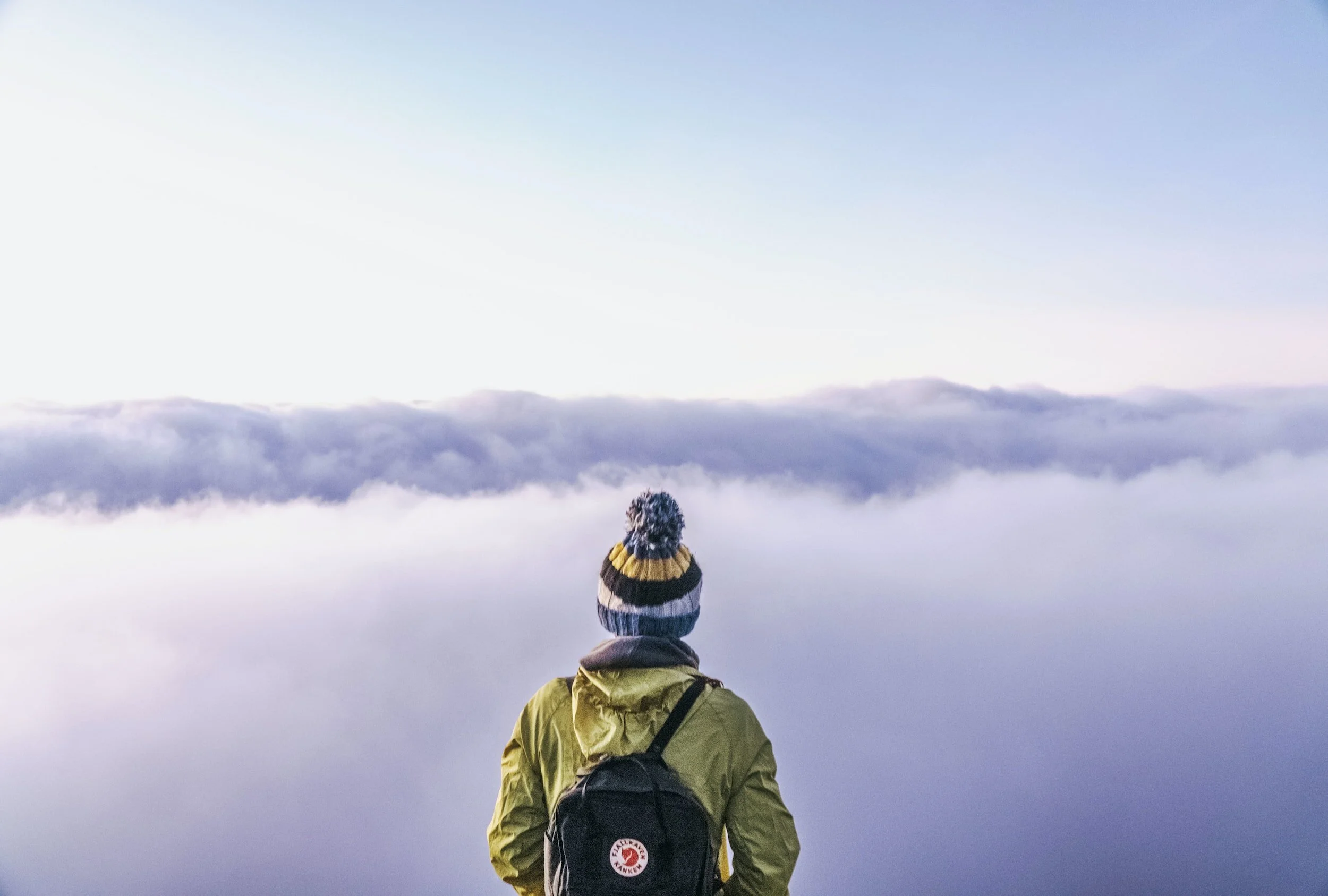 A person with a backpack and a winter hat looking at a mountain or cloud-covered landscape during dawn or dusk.