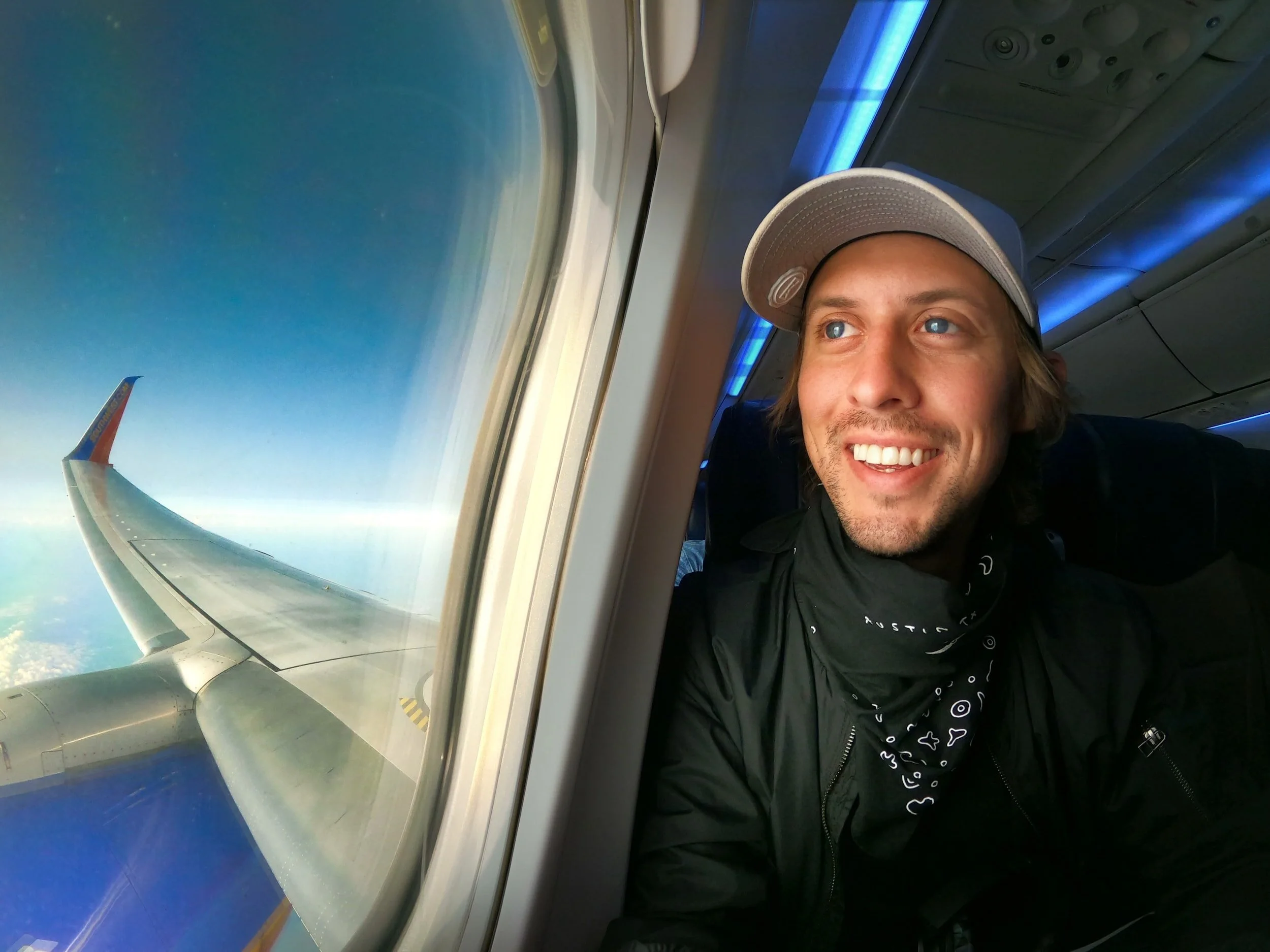 A man with a grey cap smiling while sitting by an airplane window, with a view of the wing and the sky outside.