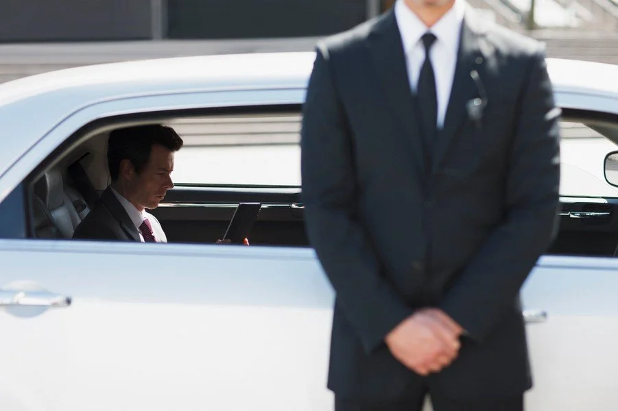 Man in a business suit sitting in a white car and looking at a tablet, while another man in a suit stands outside the car with his hands clasped in front of him.
