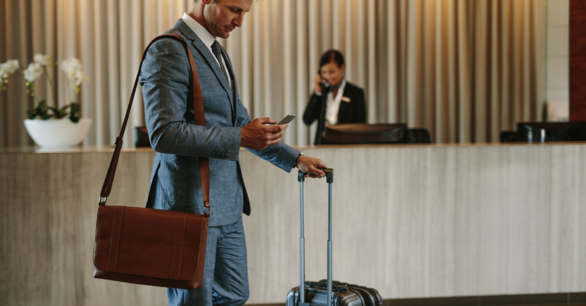 Businessman at a hotel check-in counter looking at his phone with a rolling suitcase, while a receptionist talks on the phone in the background.