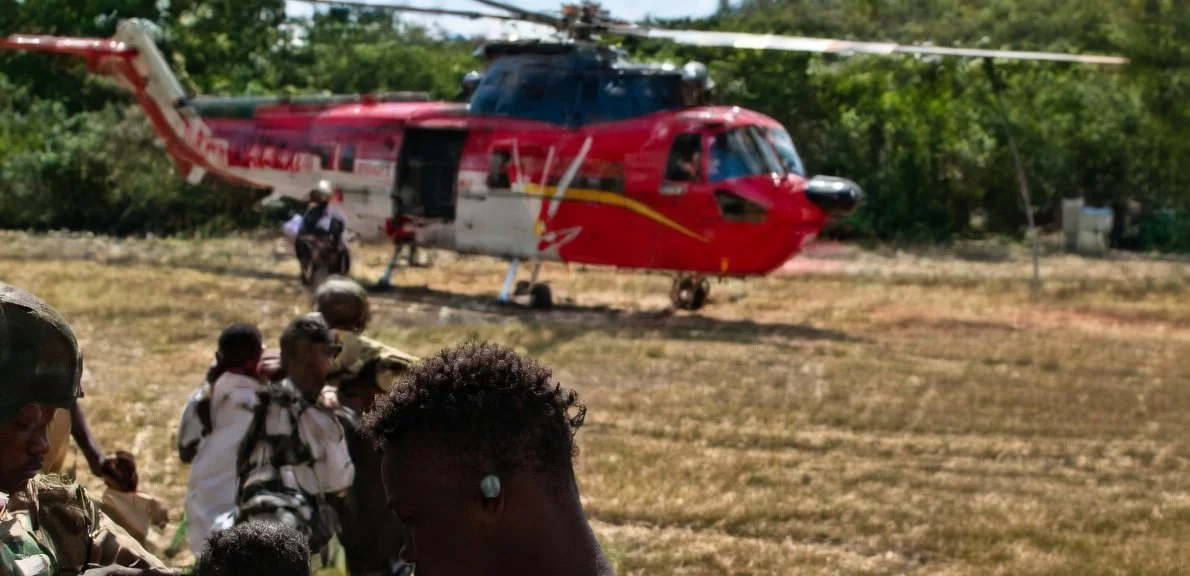 Group of people in military or paramilitary uniforms walking on a grassy field with a red and white helicopter in the background.