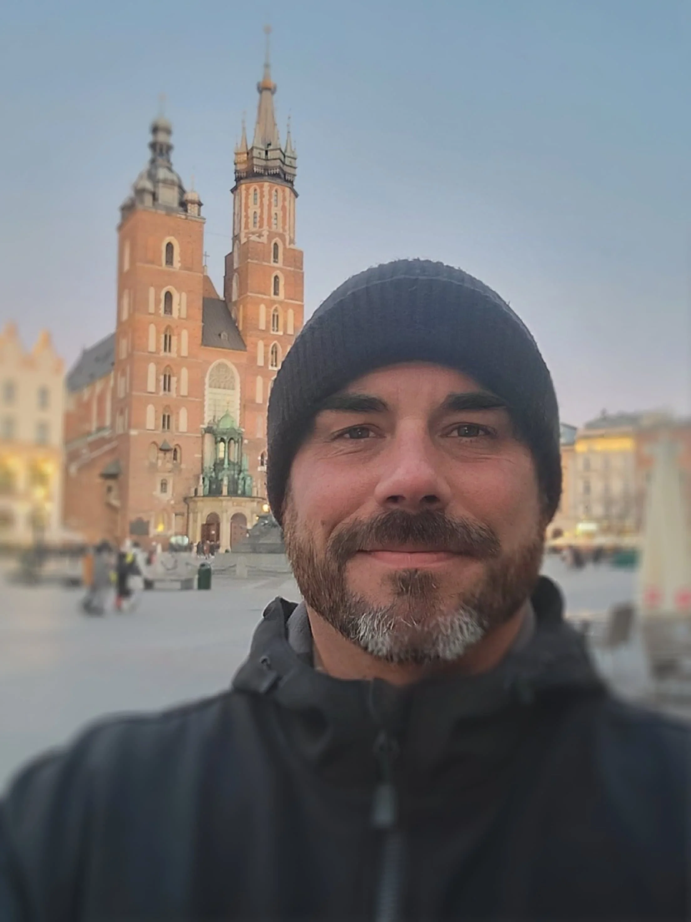 Travel Security Consultant in a black beanie smiles in front of a large cathedral with two towers in a city square at dusk.
