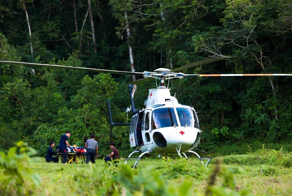 A yellow medical helicopter parked on a red running track next to an emergency response vehicle with markings for civil protection, with a cloudy sky overhead and residential buildings in the background.