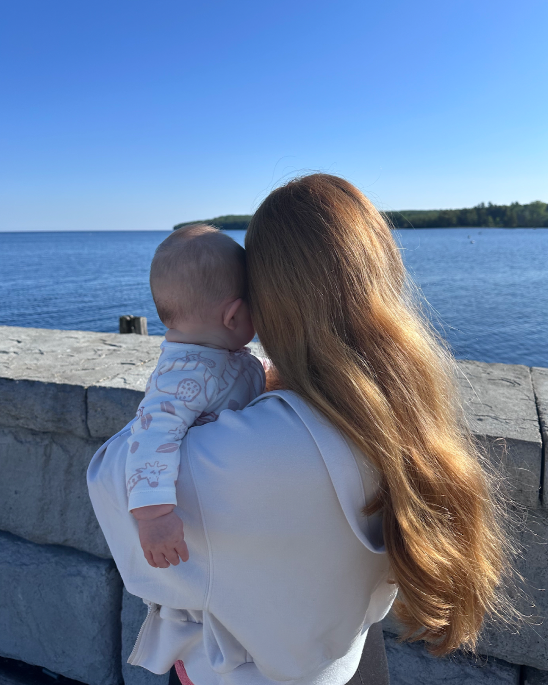 A woman with long red hair holding a baby by a stone wall at a waterfront during a sunny day with clear blue sky.