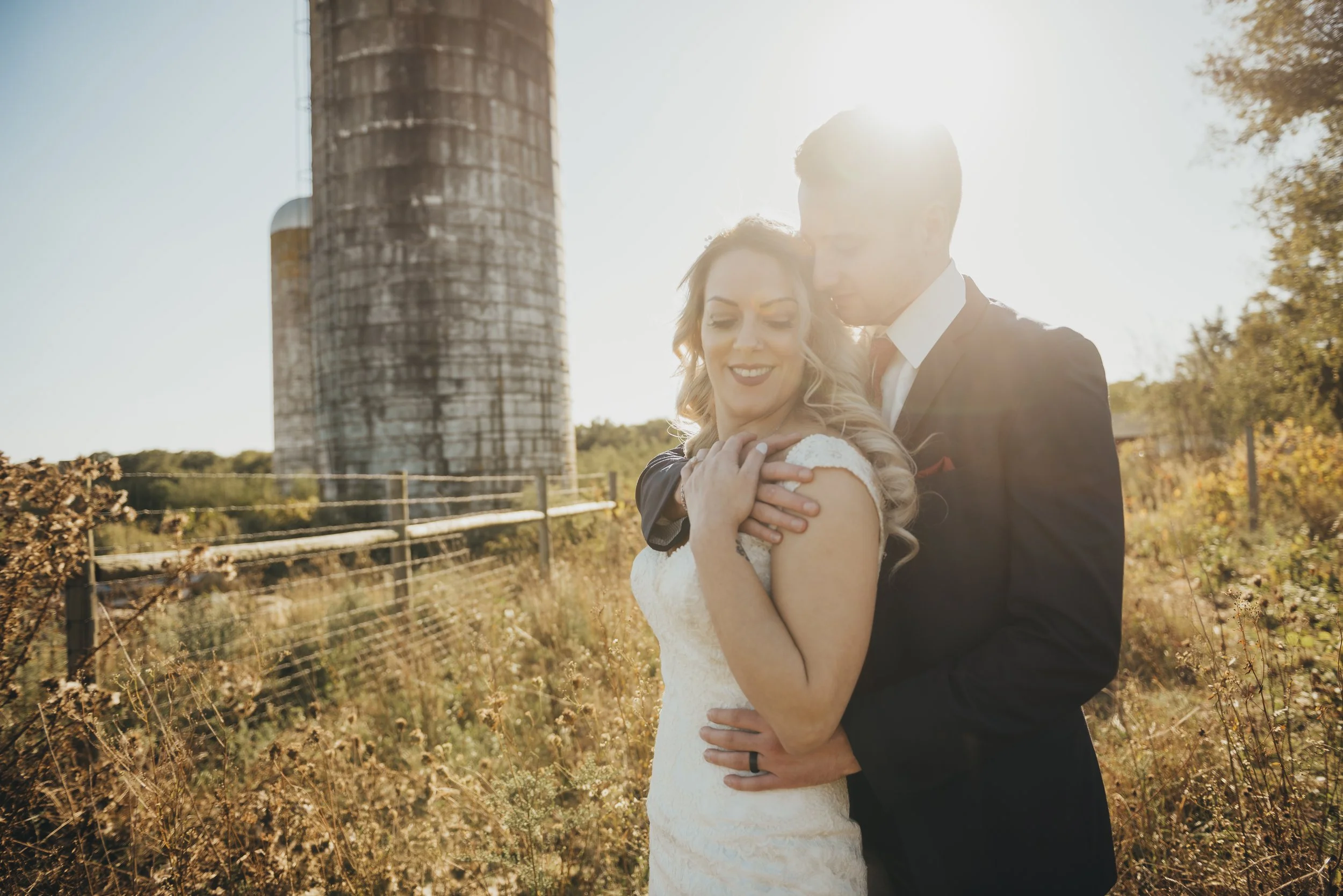 A newlywed couple embracing outdoors during sunset near an old silo and rustic fence, with golden light illuminating their faces.