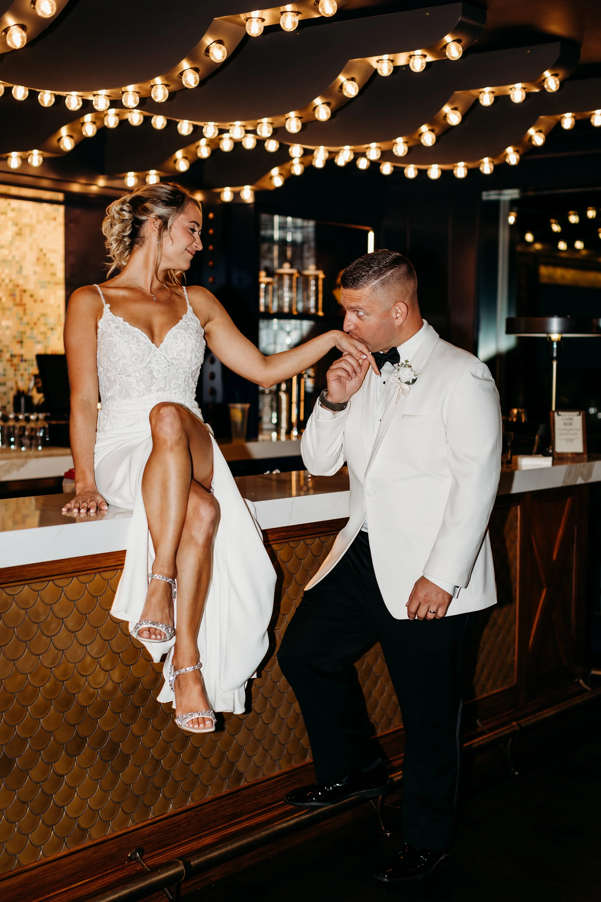 A bride and groom share a romantic moment at a bar during their wedding reception. The bride is sitting on the bar countertop, playfully touching the groom's lips. She is wearing a white wedding dress with lace details and high-heeled sandals. The gr