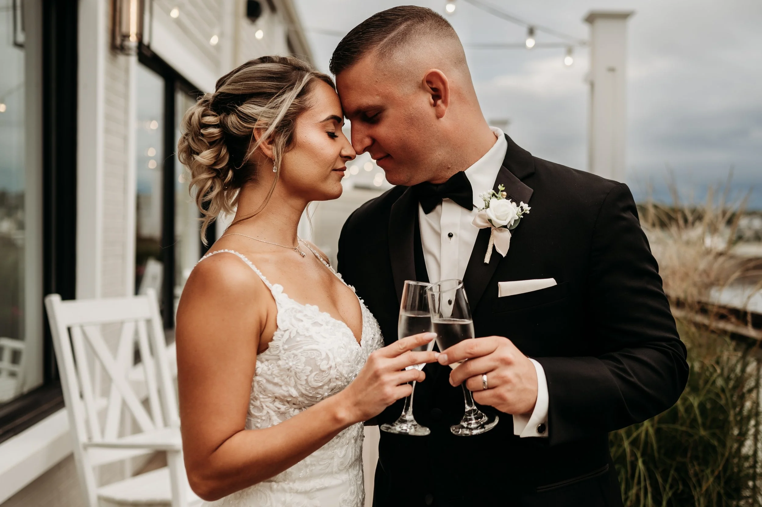 Bride and groom toasting with champagne glasses during their wedding, touching foreheads with eyes closed, on a balcony outdoor setting.