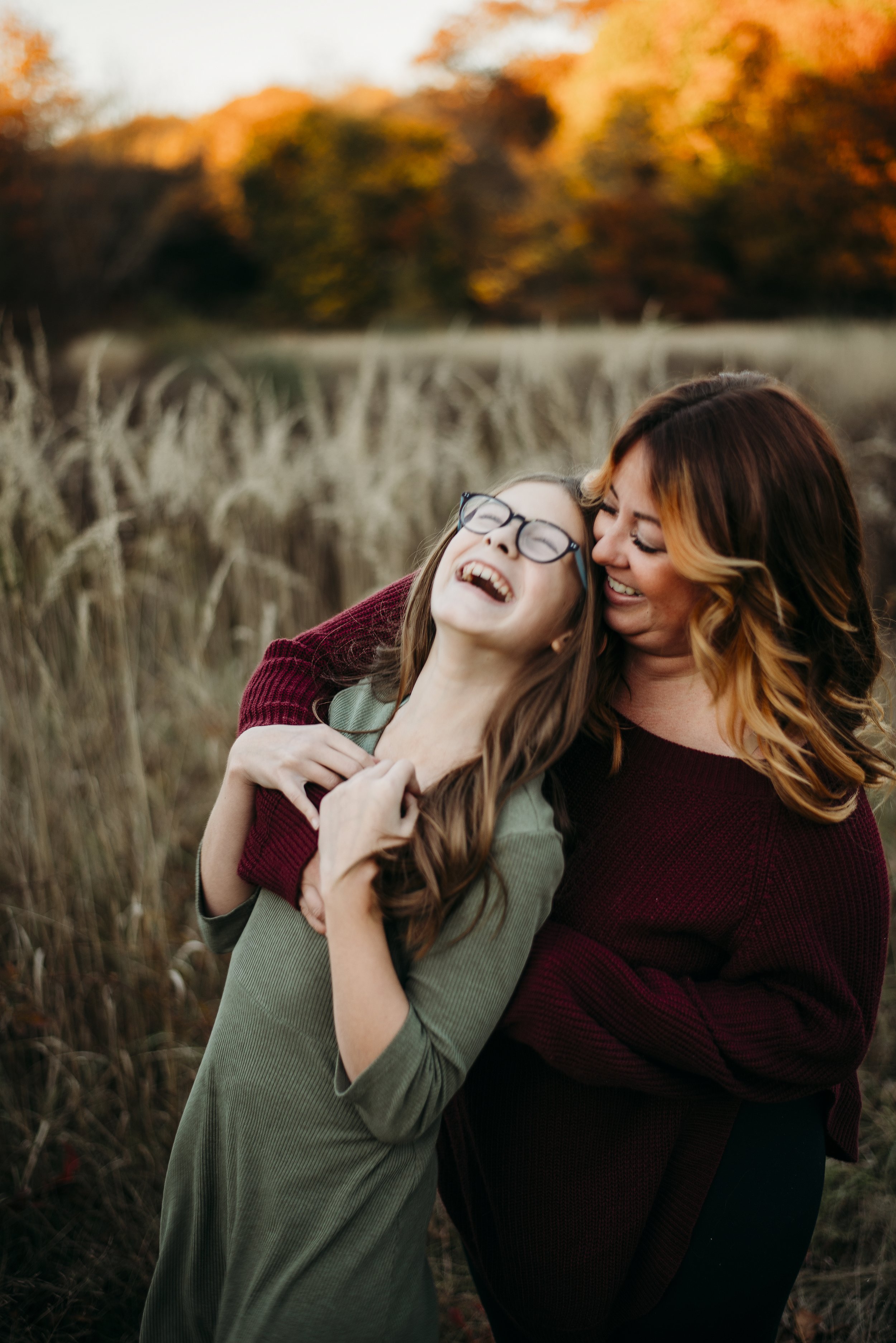 A woman and a girl sharing a joyful moment outdoors in a field during autumn, with colorful fall trees in the background.