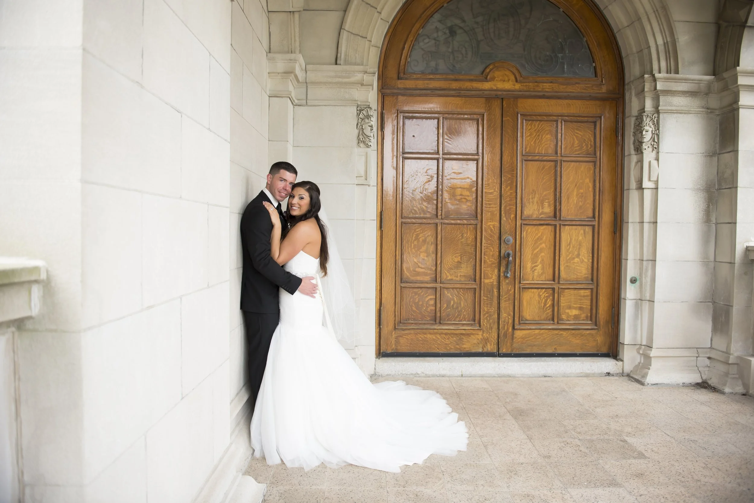 A bride and groom are smiling and hugging near a large wooden door on a stone building exterior.