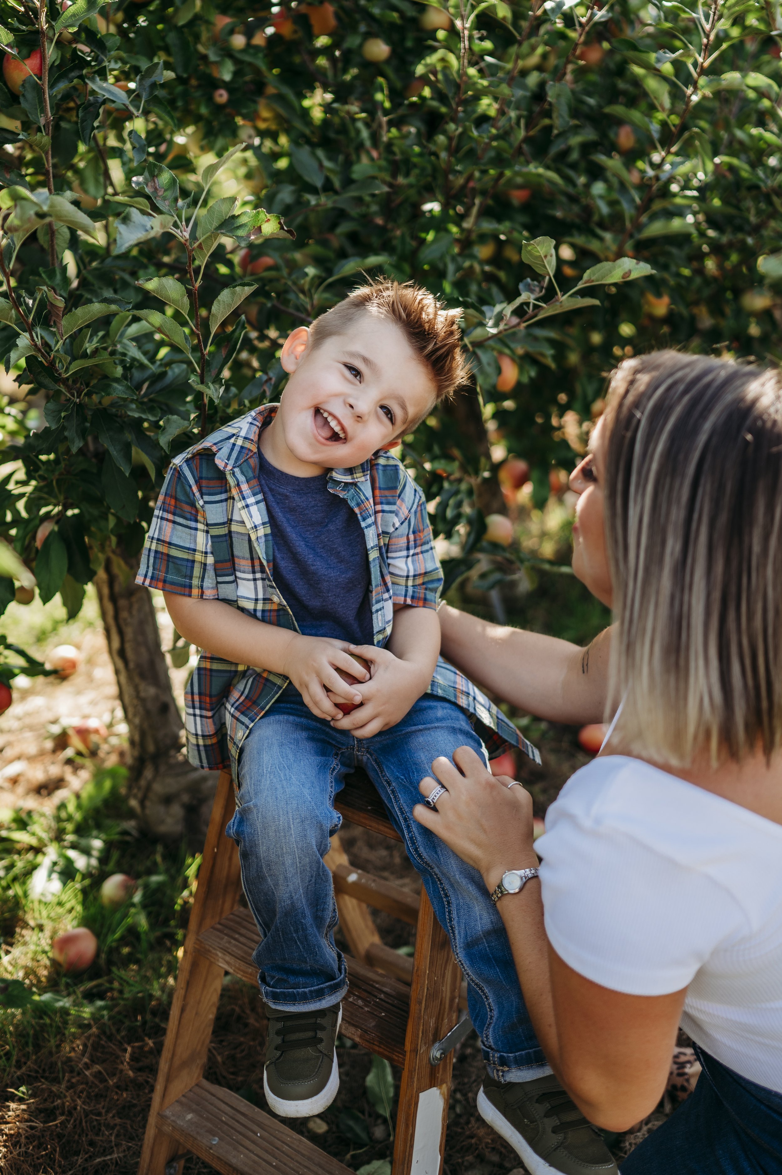 A young boy sitting on a small wooden step stool outdoors, smiling and leaning toward a woman next to him among apple trees with ripe apples, on a sunny day.